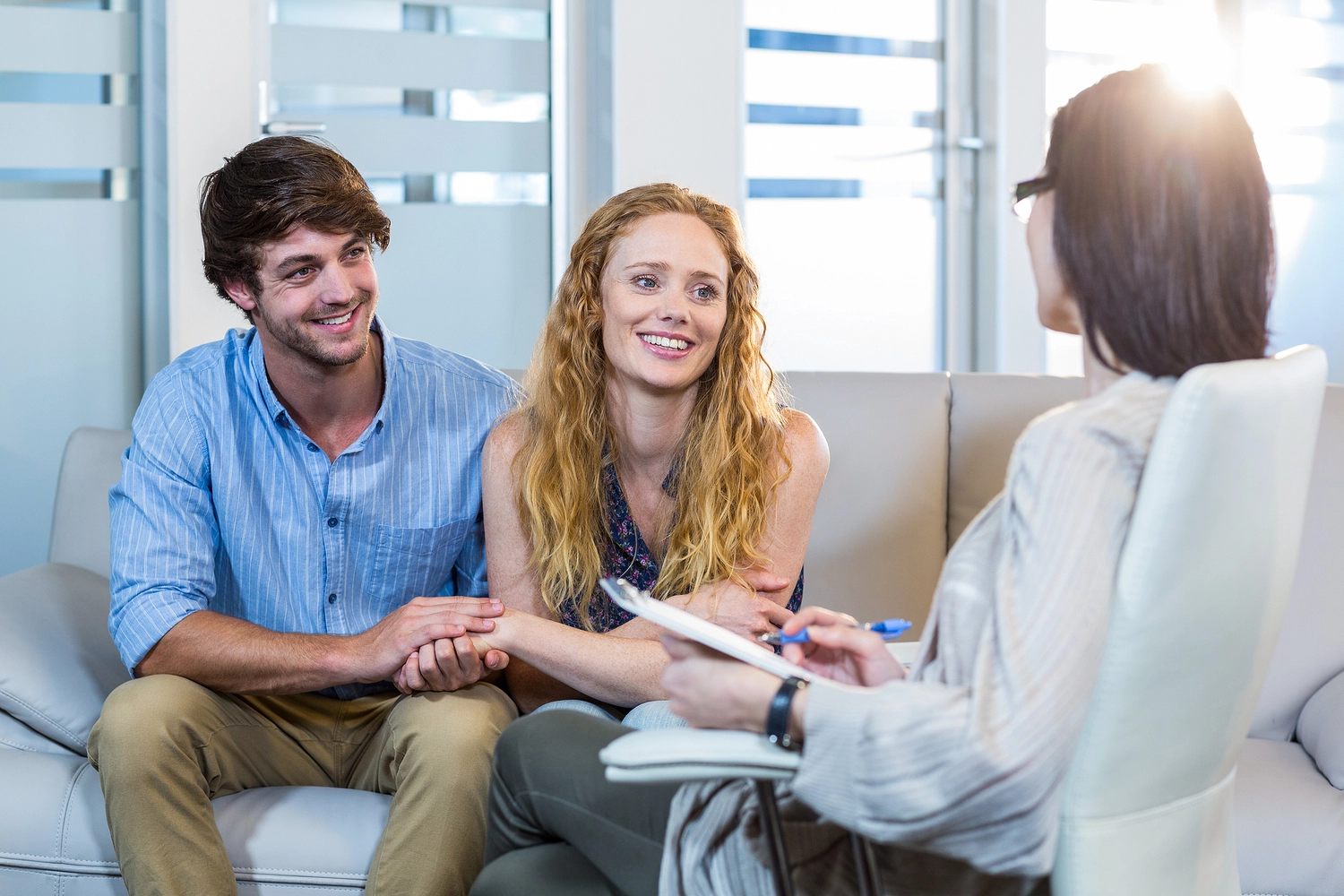 A couple sits on a sofa in a counseling session, holding hands while a therapist takes notes and engages with them. A couple sits on a sofa in a counseling session, holding hands while a therapist takes notes and engages with them