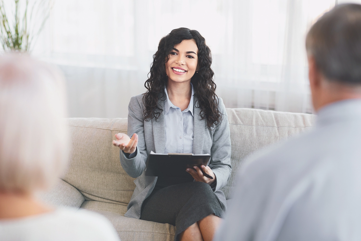 A professional woman in a suit gestures while holding a clipboard, engaged in a discussion with two clients in a cozy setting